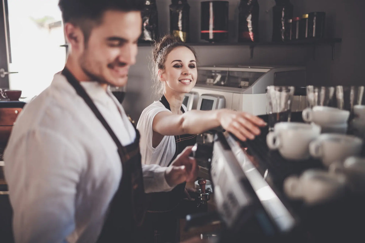 Two smiling baristas in hospitality uniforms online working at a coffee shop