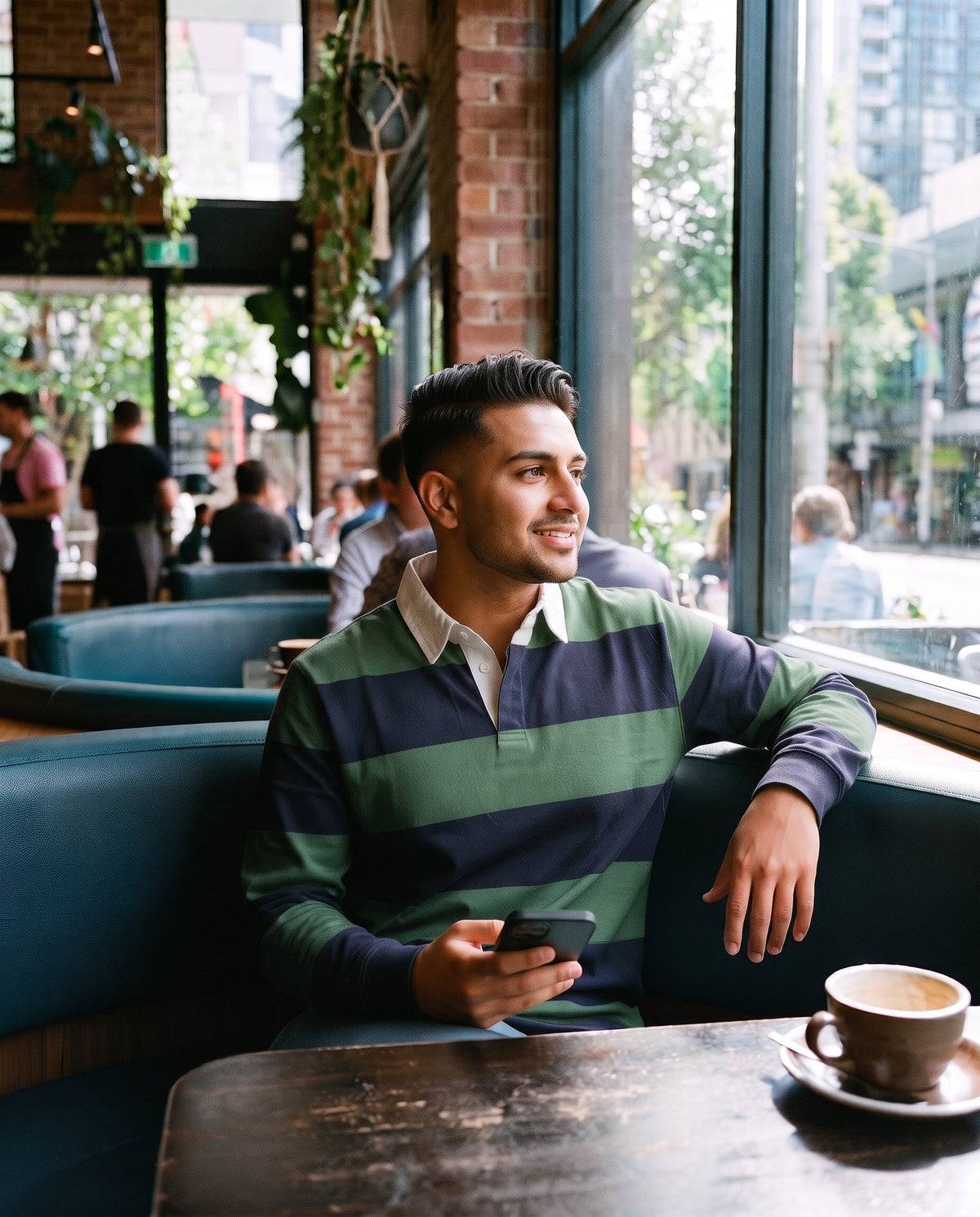A man sitting in a cozy cafe booth, wearing a green and navy striped long-sleeve rugby shirt, holding a black smartphone. A cup of coffee sits on the table in the foreground, with brick walls, teal booths, and other cafe patrons visible in the background.