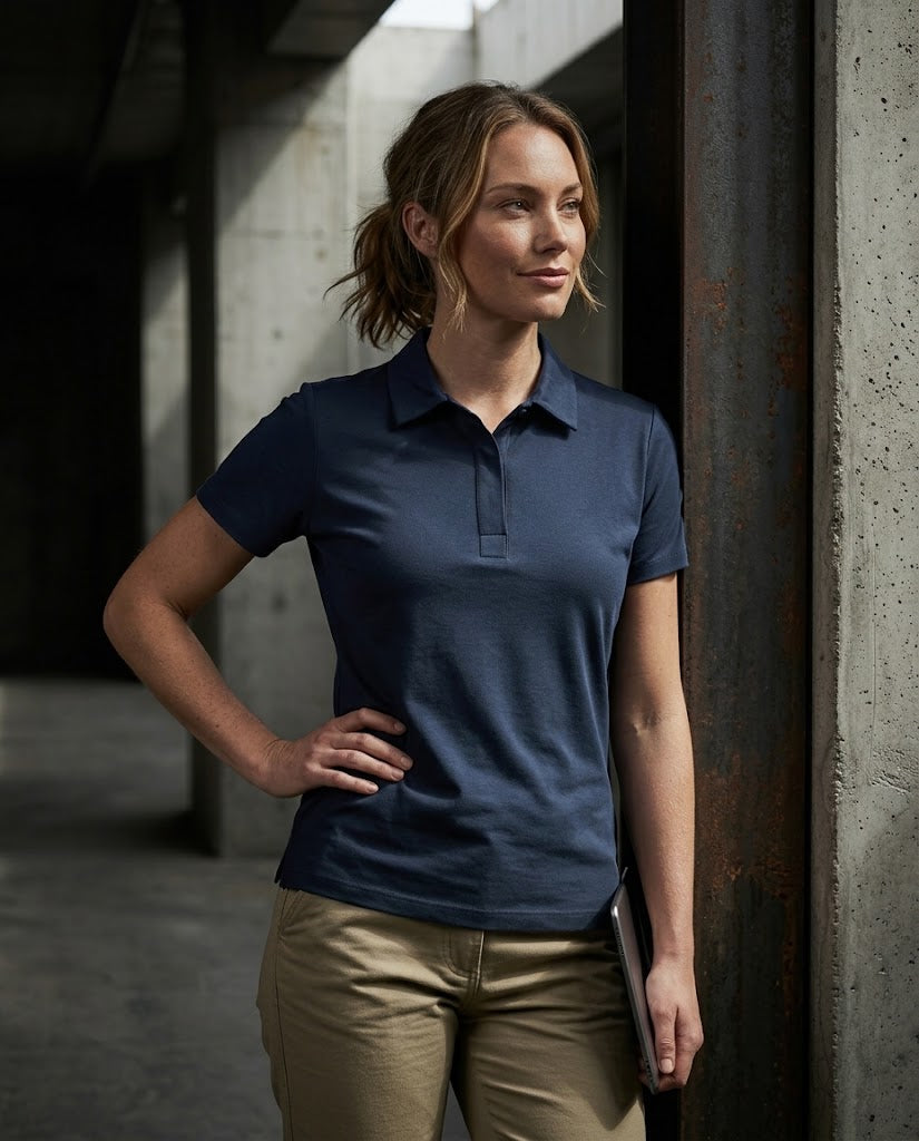 A woman wearing a navy blue short-sleeve polo shirt and khaki pants, standing in a modern industrial setting with concrete pillars.