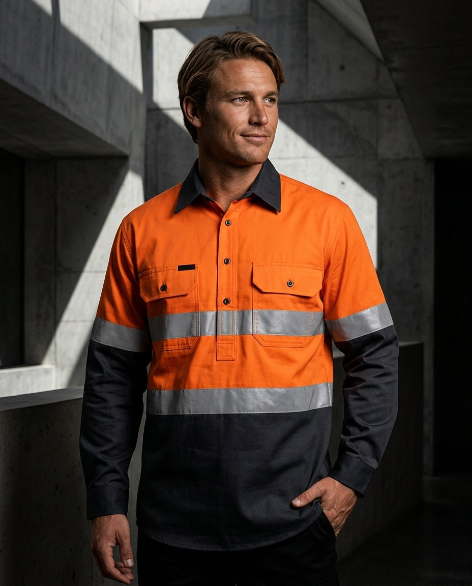 A man wearing a high-visibility orange and dark gray long-sleeve work shirt with reflective silver stripes, standing in a concrete structure with dramatic shadows.
