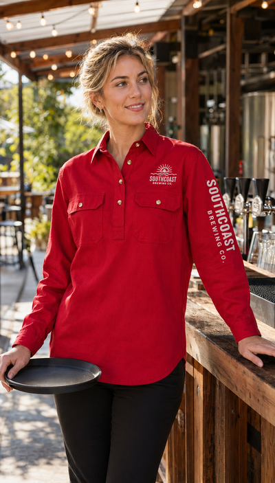 A woman wearing a red long-sleeve button-up shirt with 'SOUTHEASTERN CO.' and a logo on the chest and sleeve, standing at an outdoor bar counter and holding a black tray.