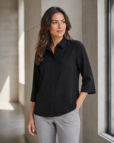 A woman wearing a black button-down shirt with three-quarter sleeves and light gray trousers, standing in a modern concrete corridor.