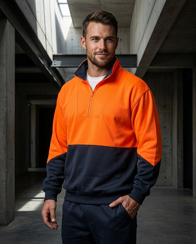 A man wearing a two-tone high-visibility quarter-zip sweatshirt in bright orange and navy blue, paired with matching navy pants, standing in a concrete industrial corridor.