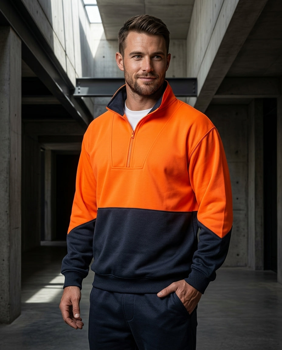 A man wearing a two-tone high-visibility quarter-zip sweatshirt in bright orange and navy blue, paired with matching navy pants, standing in a concrete industrial corridor.