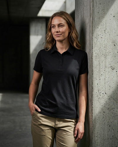 A woman wearing a black short-sleeve polo shirt and khaki pants, leaning against a concrete pillar in a modern architectural setting.