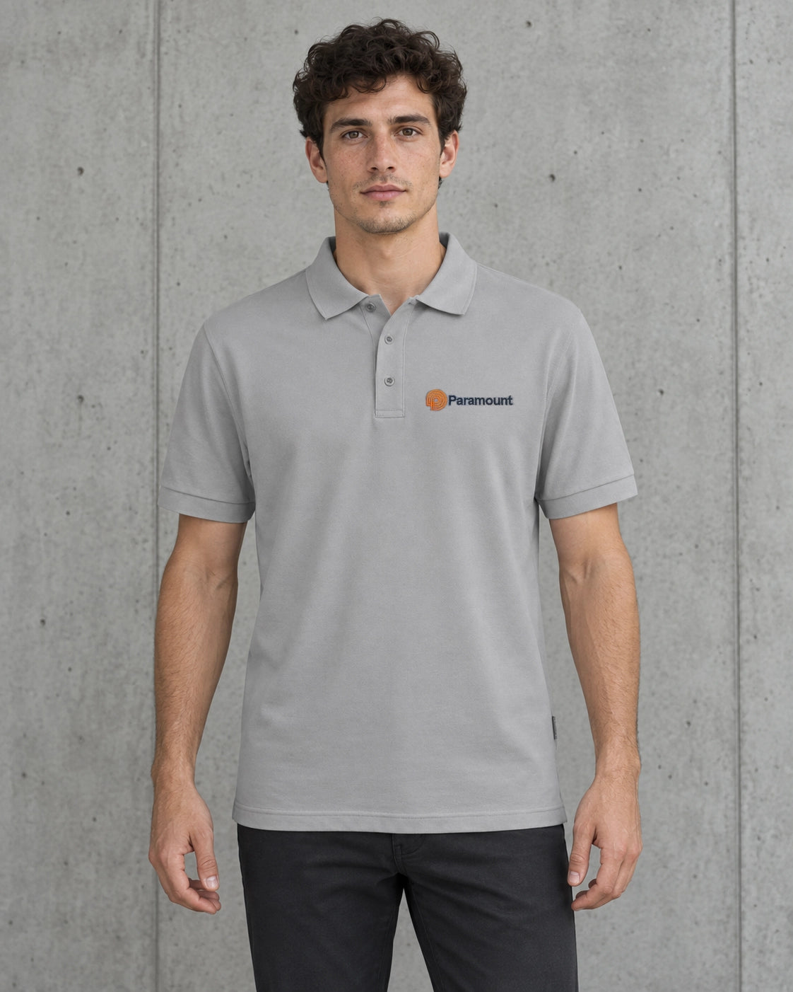 A man wearing a light gray short-sleeve polo shirt with a small orange and black Paramount logo on the left chest, standing against a textured gray concrete wall.