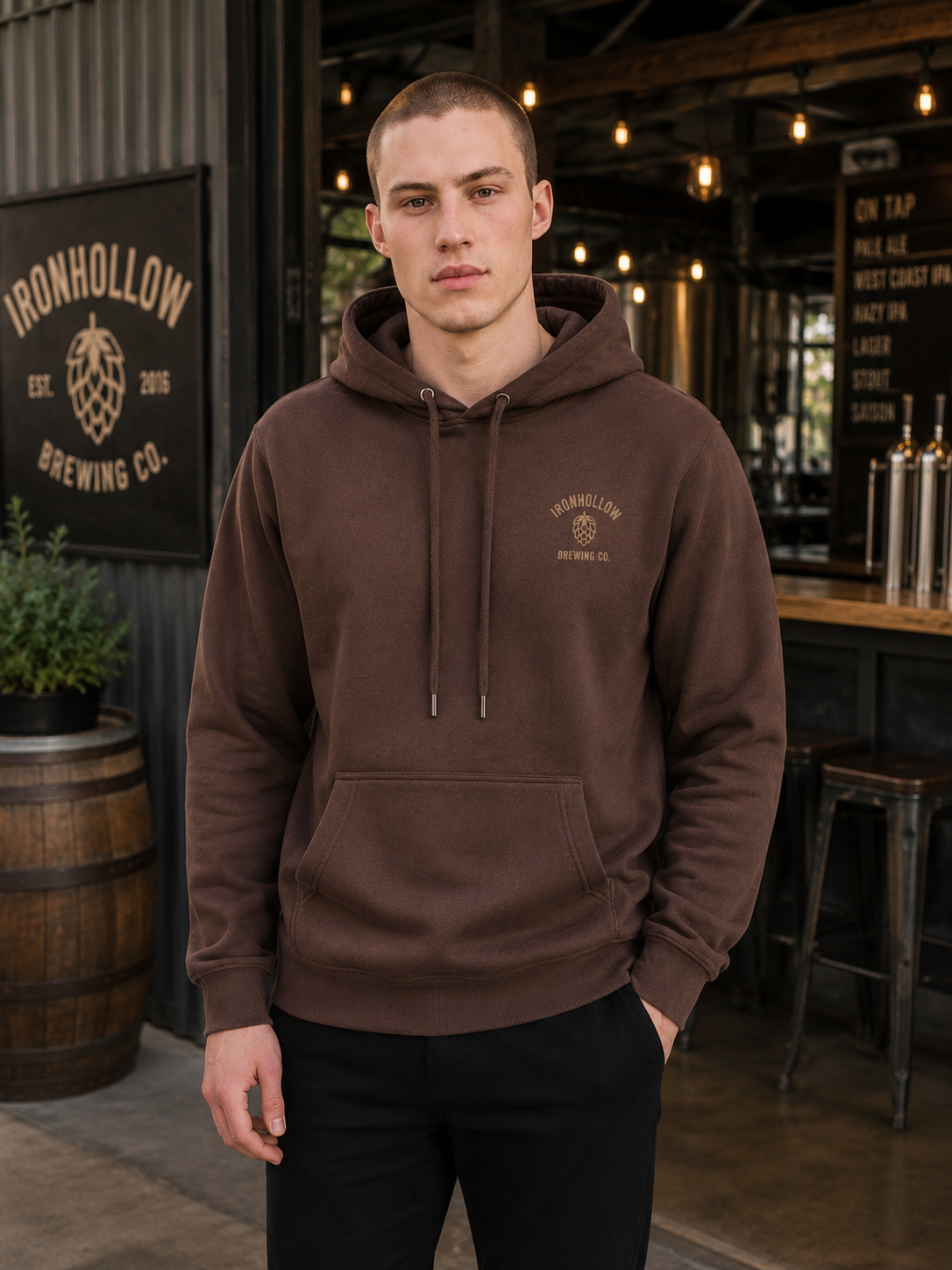 A man wearing a brown pullover hoodie with a drawstring hood and a kangaroo pocket, standing in a brewery setting. The hoodie features a small embroidered 'Ironhollow Brewing Co.